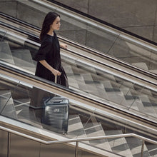 Load image into Gallery viewer, A young woman wearing a black dress heading down on an escalator with her navy blue suitcase.