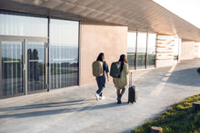 Load image into Gallery viewer, Two young women carrying their Thule backpacks and a suitcase while walking next to a building.