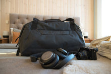 Load image into Gallery viewer, The black duffle arranged on top of a bed with a pair of headphones and a camera.