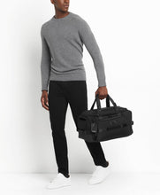 Load image into Gallery viewer, A young man holding the black duffel bag by the handle straps. Set on a white background.