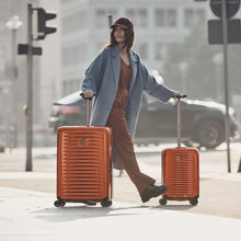 Load image into Gallery viewer, A young woman rolling two orange Victorinox suitcases in an urban setting.
