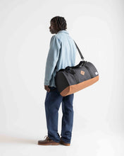 Load image into Gallery viewer, A young man carrying the black and tan duffle bag across his torso with the adjustable shoulder strap.
