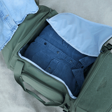 Load image into Gallery viewer, A pair of hands pulling clothes out from the moss green bag's main compartments. 
