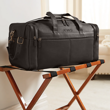 Load image into Gallery viewer, Angled view of the brown leather duffle bag set on a foldable wooden stand in a hotel setting.
