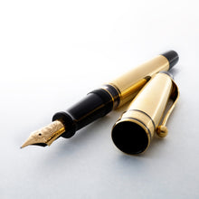 Load image into Gallery viewer, Arrangement of the fountain pen with the cap set to the side. The pen&#39;s barrel and cap are made of 18k gold, with black resin trims on the grip section and the top of the pen. 
