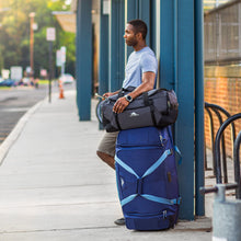 Load image into Gallery viewer, A man in an outdoor urban area standing next to a blue wheeled duffle with the black Fairlead travel duffle set on top.
