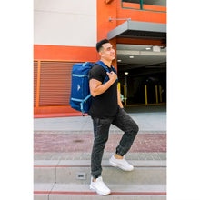 Load image into Gallery viewer, A young man in an outside urban setting carrying the navy blue duffle backpack.
