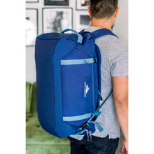 Load image into Gallery viewer, A young man facing forward in a blurry background, carrying the navy blue duffle backpack on his back.
