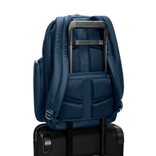 Load image into Gallery viewer, Angled back panel view of the navy blue backpack with the attachment feature holding it to a suitcase.
