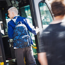 Load image into Gallery viewer, A young man carrying the Cheetah Camo backpack steps into a school bus. 
