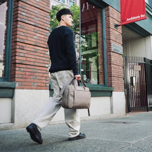 Load image into Gallery viewer, A young man walking on a sidewalk while carrying the light brown Gibson bag by the top grip handles. 
