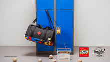 Load image into Gallery viewer, Black duffel bag with colorful accents hanging on a blue locker, with a radio, with Lego and Herschel logos in the foreground.
