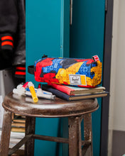Load image into Gallery viewer, Colorful pencil case with Herschel logo on a stool against a teal locker.
