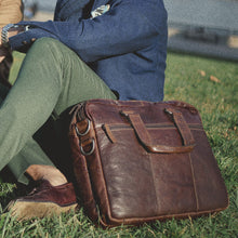 Load image into Gallery viewer, A young man wearing a suit sitting on grass with the brown leather briefcase next to him. 
