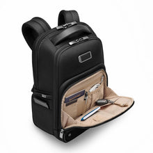 Load image into Gallery viewer, Angled front view of the black backpack with the bottom from panel pocket unzipped. A passport, USB cable, pen, notebook, and keys attached to a key sling are organized in the compartments.
