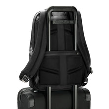 Load image into Gallery viewer, Back panel view of the black backpack with the horizontal attachment strap holding it onto the extended handle of a suitcase.
