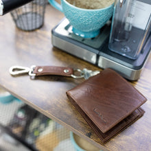 Load image into Gallery viewer, The cognac leather wallet on a wooden table next to an espresso machine.
