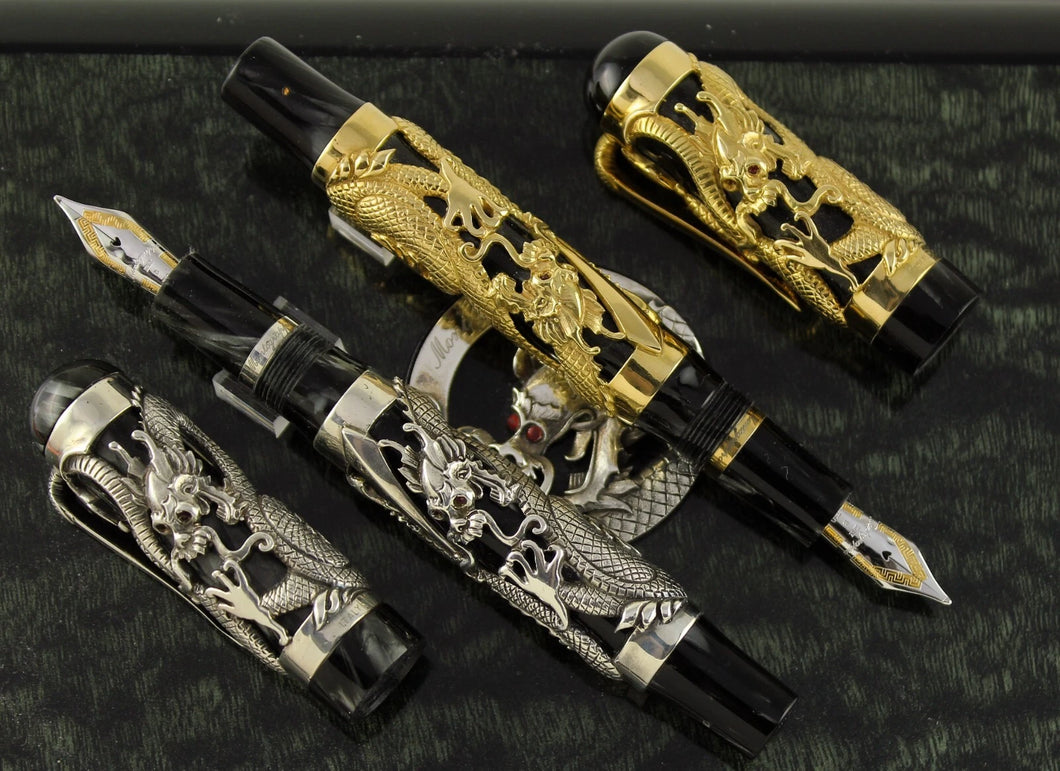 The Montegrappa 1995 Dragon fountain pens, uncapped, in gold and silver overlay, featuring intricate dragon engravings on black marbled celluloid bodies, displayed on a dark background.
