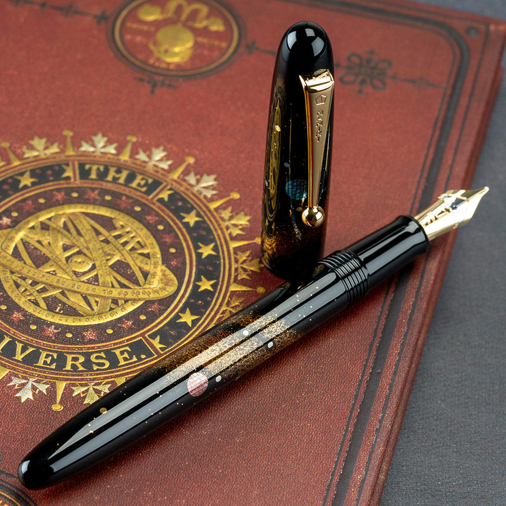 Angled view of the Fountain Pen. The cap is set off and standing on the left side. It is placed on top of a red book cover featuring an elaborate decoration with an Astrology design. It reads: 