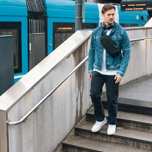 Load image into Gallery viewer, Man in a denim jacket and black pants walking downstairs while carrying the sling bag across his torso.
