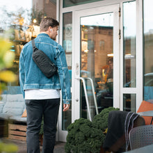 Load image into Gallery viewer, Man in an urban setting, carrying the black sling bag on his back. 
