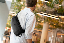 Load image into Gallery viewer, A young man wearing the sling bag across his torso on a blurred urban setting. 
