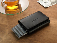 Angled view of the Pularys Boston wallet in black on a brown wooden table. There is a glass tea cup and a $20 bill next to it.