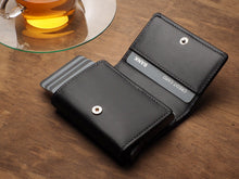Load image into Gallery viewer, Angled view of the opened Pularys Boston wallet in black on a brown wooden table. A glass tea cup is behind the wallet.  
