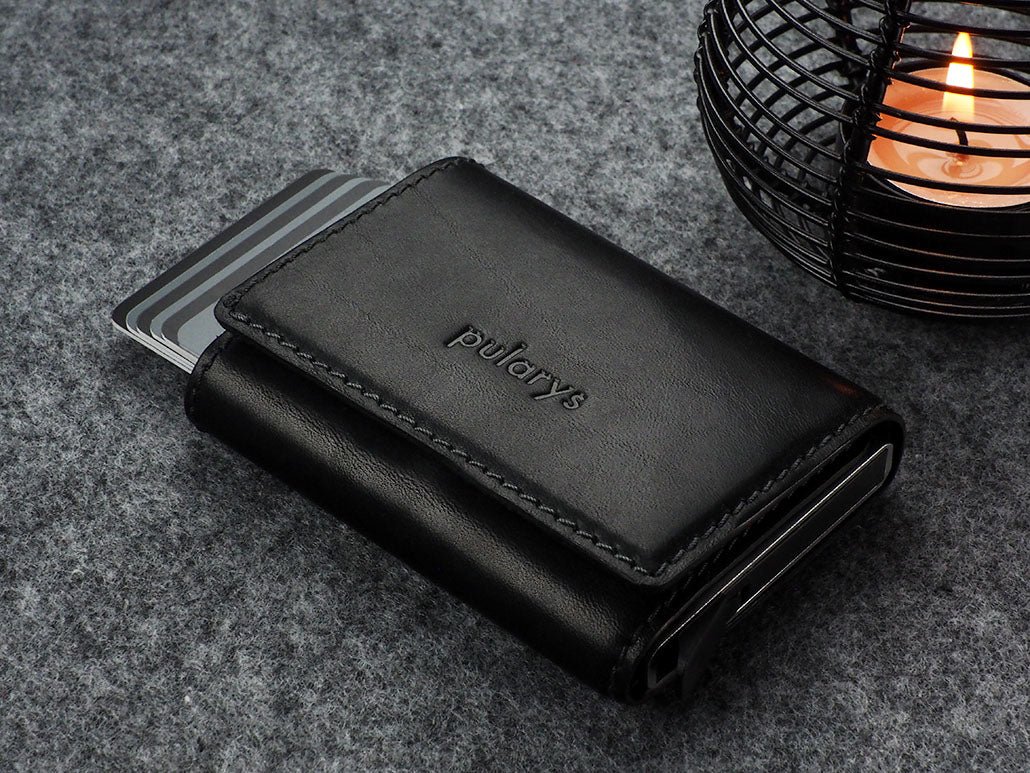 Angled view of the closed Pularys Colorado RFID black leather wallet. Set on a gray textured surface with a candle set next to it.