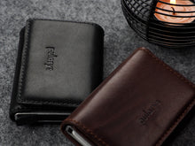 Load image into Gallery viewer, Angled view of the closed Pularys Colorado RFID leather wallets in black and brown. Set on a gray textured surface with a candle set next to it.
