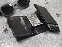 Load image into Gallery viewer, A black leather-covered aluminum wallet arranged with a ballpoint pen, alongside sunglasses, jax  and money.
