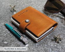 Load image into Gallery viewer, Angled view of the cognac wallet arranged with a pen, sunglasses and jax toys. 
