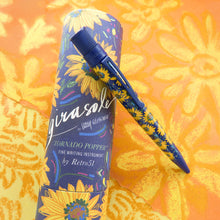 Load image into Gallery viewer, The pen leaning on the matching gift tube. Set on an orange background with yellow flowers.

