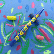 Load image into Gallery viewer, Side view of the pen on a blue background with some oil pastel pieces and hand drawn flowers.
