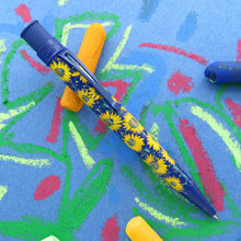 Load image into Gallery viewer, View of the pen leaning on a yellow oil pastel. Set on a blue background with sketches drawn on it.
