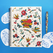 Load image into Gallery viewer, The sketchbook arranged with the pen and some tattoo designs on a blue background. 
