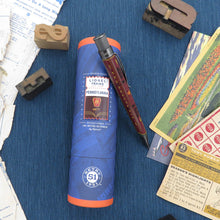 Load image into Gallery viewer, Pen and blue commemorative tube on a textured surface with vintage items.
