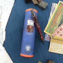 Load image into Gallery viewer, Pen and blue commemorative tube on a textured surface with vintage items.
