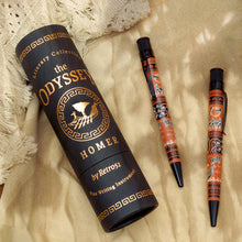 Load image into Gallery viewer, The black and gold color presentation tube, next to two of the rollerball pens on a sandy background.
