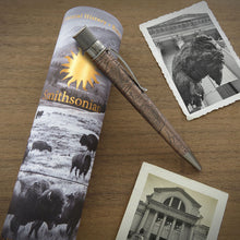 Load image into Gallery viewer, The acid-etched bison pen with a brushed brass clip and tip lies across a rolled Smithsonian-themed printed presentation tube featuring bison imagery, placed on a wooden surface with two black-and-white photo prints nearby.
