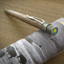 Load image into Gallery viewer, The rollerball pen with an acid-etched Bison on the barrel pattern and a brushed brass clip, resting on top of its presentation tube over a wooden tabletop.
