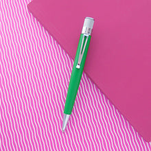Load image into Gallery viewer, A green retractable pen with silver accents lies diagonally across a bright pink background with wavy white lines and a solid pink notebook.

