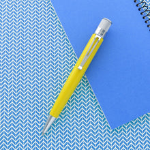 Load image into Gallery viewer, A yellow pen with chrome accents lies diagonally across a blue patterned surface and a blue notepad.
