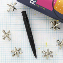 Load image into Gallery viewer, Black Stealth ballpoint on a grid surface with decorative objects around it.
