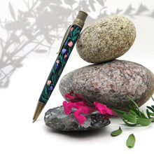Load image into Gallery viewer, Side view of the Growth pen leaning on three decorative stones with flowers.

