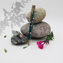 Load image into Gallery viewer, Side view of the Growth pen leaning on three decorative stones with flowers.
