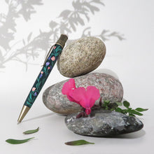 Load image into Gallery viewer, Side view of the Growth pen leaning on three decorative stones with flowers.
