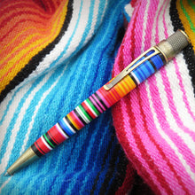 Load image into Gallery viewer, A close-up of the pen with a colorful striped sarape pattern, resting on a vibrant, multicolored knit fabric background.
