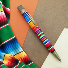 Load image into Gallery viewer, The sarape style rollerball with brushed brass accents on a brown and orange background with a colorful sarape on the side.
