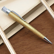 Load image into Gallery viewer, A gold color ballpoint pen with an ornate floral engraving on its barrel, a chrome clip, and a silver textured top, resting diagonally on a wooden surface next to a notebook.
